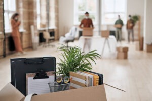 Focus on open cardboard box with office supplies, clipboards and plant against people moving into new office in background after a corporate relocation move